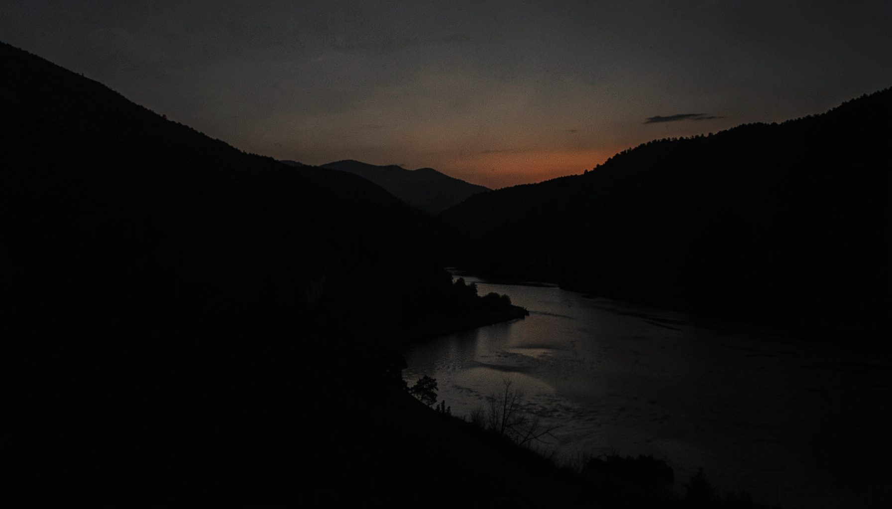 A dark, low-key photograph with deep shadow, showing the muted silhouette of a river valley at dusk in charcoal and amber tones